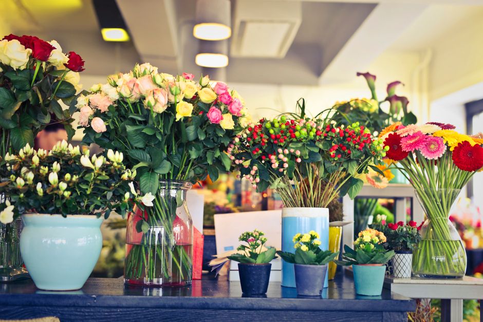 A variety of fresh flower arrangements displayed on a table, including large bouquets of pink and white roses in clear glass and ceramic vases, complemented by smaller potted yellow and pink flowers. 