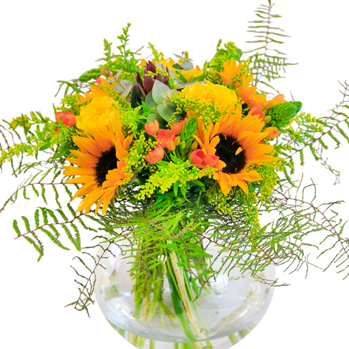 From the first glance, this bouquet is a sunlit celebration-radiant sunflowers and luxurious yellow garden roses aglow in a modern glass vase, reflecting the lively rhythms of Poplar's community. The sunflowers, with their robust golden petals sweeping outward from bold, chocolate-brown centers, hint at late-summer afternoons spent near All Saints Church or along the bustling streets of this vibrant East London neighbourhood. Between them, the garden roses unfurl with quiet poise, their buttery petals layered in soft spirals, whispering classic romance and the comfort of familiar places. Among the blooms, bright red Hypericum berries pop-glossy and spirited, providing cheerful contrast and depth among the deep greens. The bouquet's lush foundation of ferns, airy goldenrod with clusters of tiny yellow flowers, and slender green spikes weaves a rich, tactile narrative, suggesting both motion and tranquility. Bluish-green eucalyptus leaves flicker at the edges of the arrangement, adding fresh, aromatic notes that lend a gentle rusticity. The clear, spherical vase elegantly cradles every stem, the visible tangle beneath the water testimony to floral artistry and attention to detail. This arrangement is ideal for marking special milestones-from birthdays to anniversaries-or simply for sending sunshine and goodwill across Poplar and beyond, a true reflection of Poplar Florist's heartfelt connection to its community.