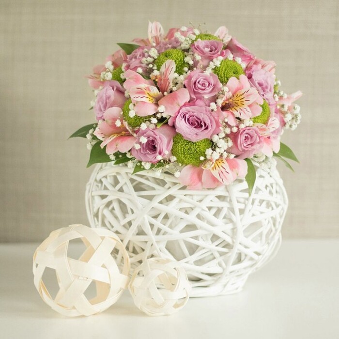 On a luminous white surface, an expertly composed floral centerpiece comes to life, radiating gentle colour and harmony-much like an elegant celebration along Poplar's Canary Wharf. The arrangement's core features full-bloomed roses in glowing pastel pink and lavender, accompanied by playful, bright green Kermit mums and delicate sprays of cloud-like white baby's breath. Graceful alstroemeria, their pale pink petals etched with subtle stripes and hints of sunshine yellow, offer a refined contrast, highlighting the nuance of expert local floristry. The bouquet is rooted in a substantial, cylindrical vase crafted from crisp white woven strips-a contemporary design that bridges modern style with hints of Poplar's traditional craft heritage. To the left, two handwoven spheres in light beige tones echo the vase's texture, providing visual balance and rustic warmth. The spheres' sizes and placement, with the larger nestled close and the smaller slightly askew, lend a dynamic yet harmonious rhythm to the composition. The soft, textured beige background draws focus to the vibrancy of the blooms and lush greenery, making the ensemble suitable for grand wedding tables, milestone celebrations, or as a distinguished centerpiece at local Poplar events. Every element-colours, shapes, placement-speaks to the attentive, creative touch offered by Poplar Florist, expressing both artistry and a deep connection to the heart of the community.
